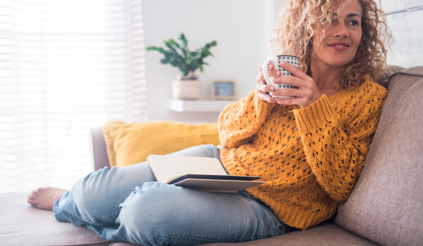 Woman relaxing on a couch with a book in her lap and holding a cup of tea, practicing self-care and prioritizing her mental health.