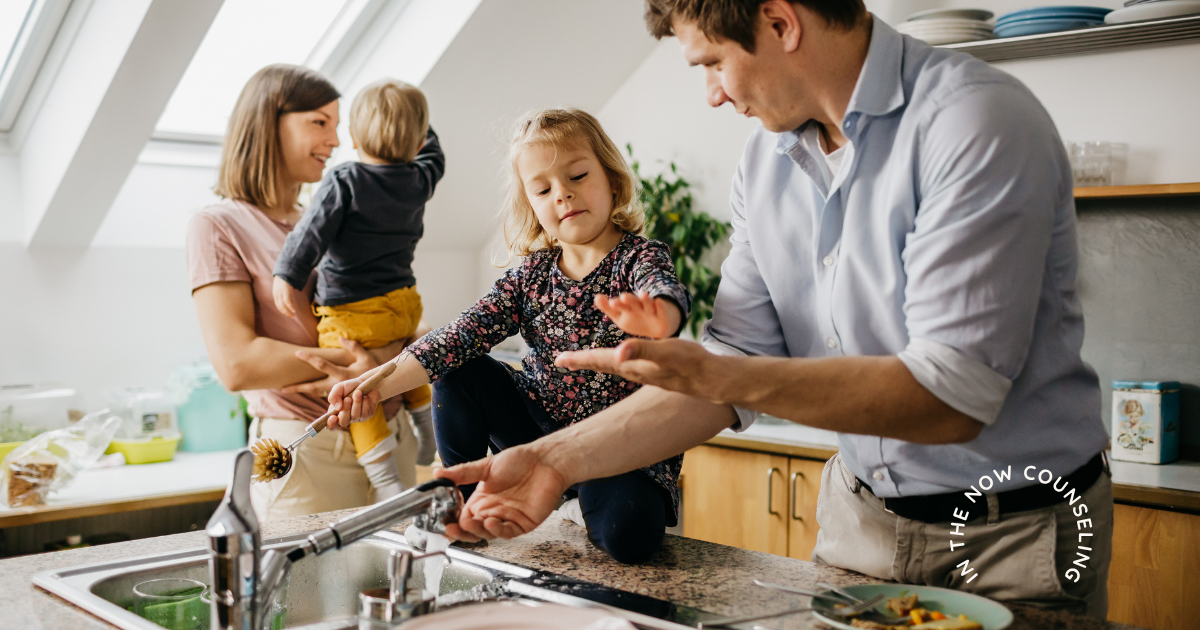 a man washing dishes with his daughter while a woman holds a baby showing how they share the mental load with their partner this summer