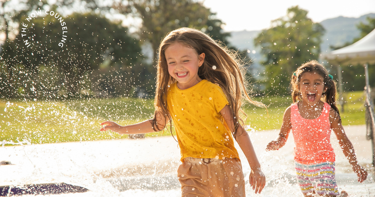 Your child's behavior in summer can look like Two young sisters happily playing in a backyard sprinkler during summer break, showing the joyful and connected moments families hope for despite summer challenges when a child's behavior can get worse in summer
